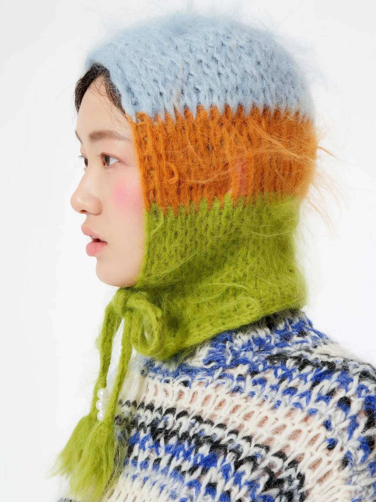A studio profile of a woman in a fuzzy mohair hood striped pale blue, orange and lime, tied under the chin, 3:4.