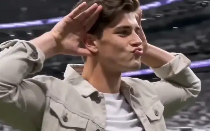 A person in a beige jacket cups both hands behind the ears in a stadium