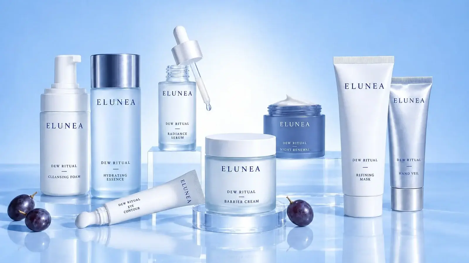 Blue studio lineup, 16:9, labeled ELUNEA DEW RITUAL: cleansing foam, hydrating essence, radiance serum with a dropper, eye contour, barrier cream, night renewal, refining mask, and hand veil, with dark grapes on a reflective surface.